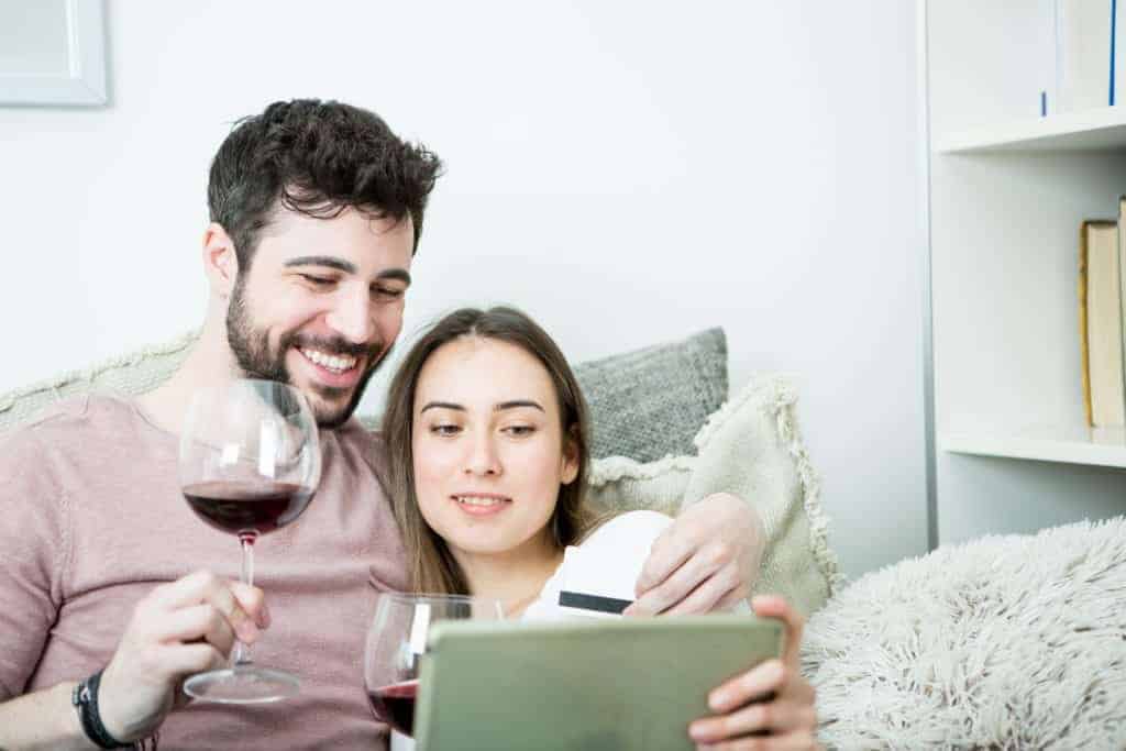 A couple sitting on their couch drinking red wine as they prepare to purchase wine online on their tablet.